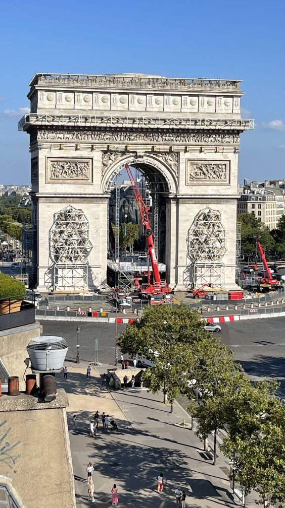 L'Arc de Triomphe à Paris est en cours de rénovation, entouré de grues et d'échafaudages, avec un ciel bleu clair au-dessus.