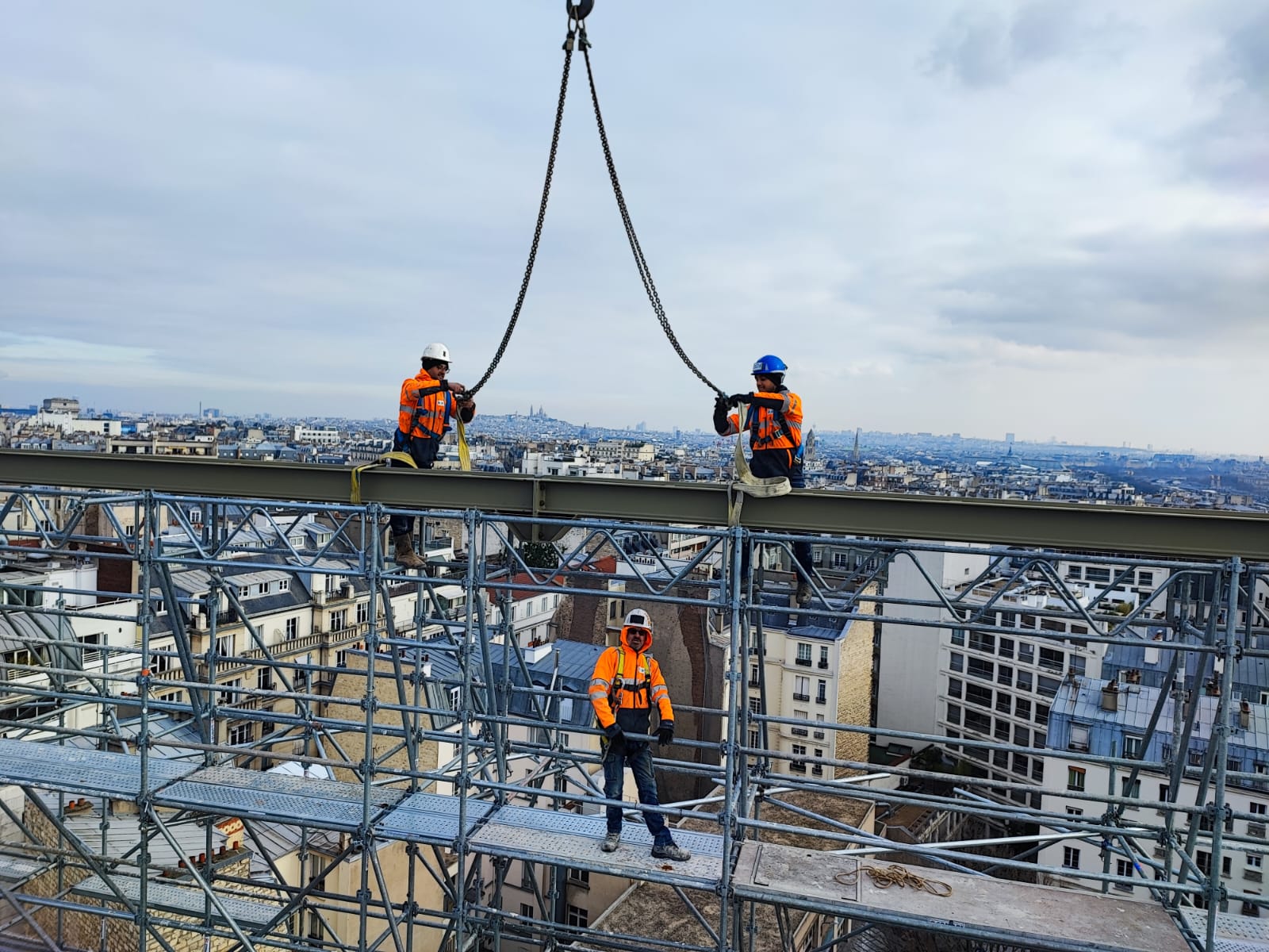 Trois ouvriers du bâtiment en tenue de sécurité naviguent sur une poutre au sommet d'une structure, avec une vue panoramique sur la ville en contrebas.
