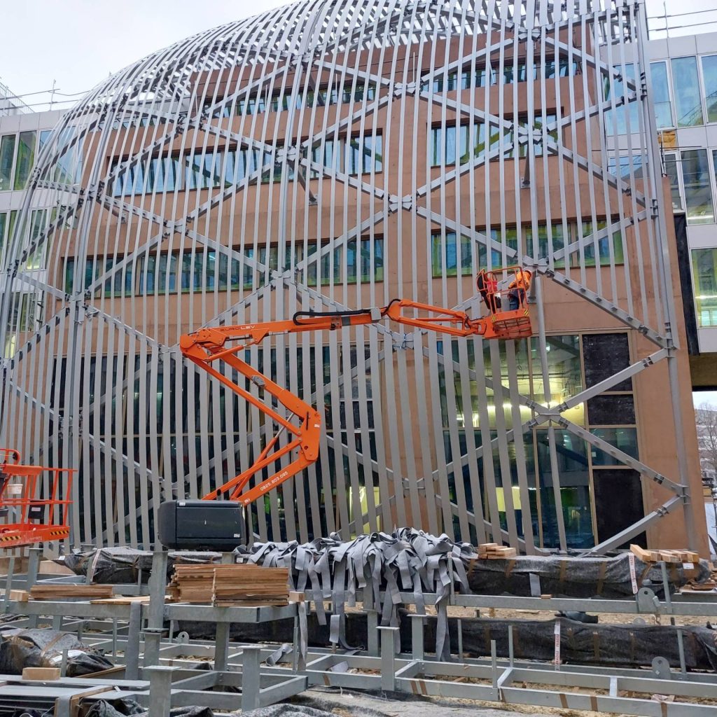 Bâtiment moderne en construction, avec sa façade métallique incurvée distinctive. Un ascenseur orange permet à un ouvrier d'ajuster la structure, créant ainsi une impression de progrès.